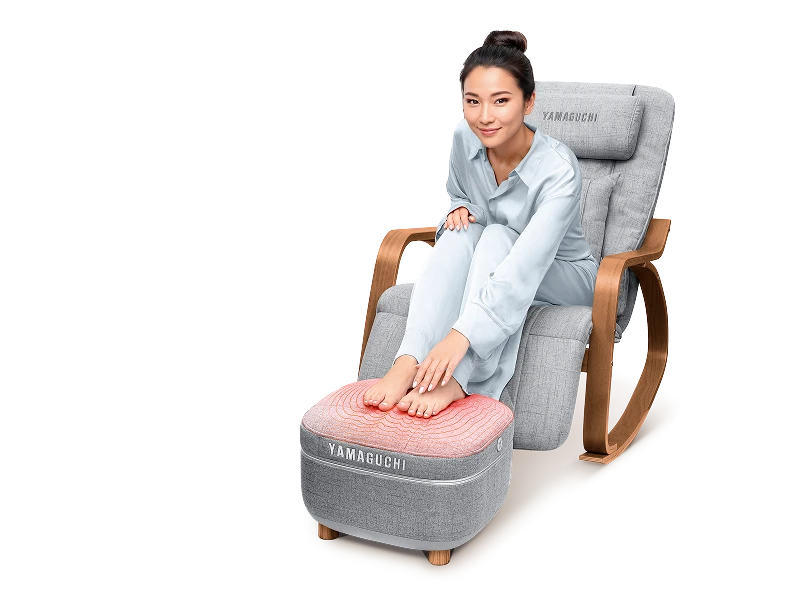 Женщина в белой пижаме сидит в массажном кресле Yamaguchi Liberty Comfort, положив ноги на Yamaguchi Capsula Grey
